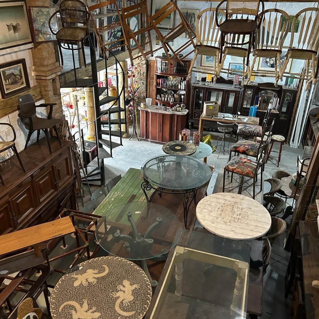 Parte interna do galpão, onde temos diversos objetos, como mesas, cadeiras, quadros e entre outros.