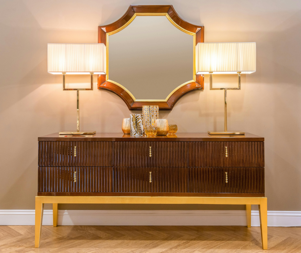 Cômoda alta em madeira escura com acabamento de alto brilho e três gavetas com padrão de frisos verticais. A base é dourada, assim como os puxadores. O móvel está centralizado entre duas luminárias com bases geométricas de metal dourado e cúpulas beges, e acima dele, um espelho de parede com moldura curva em madeira e detalhes dourados.