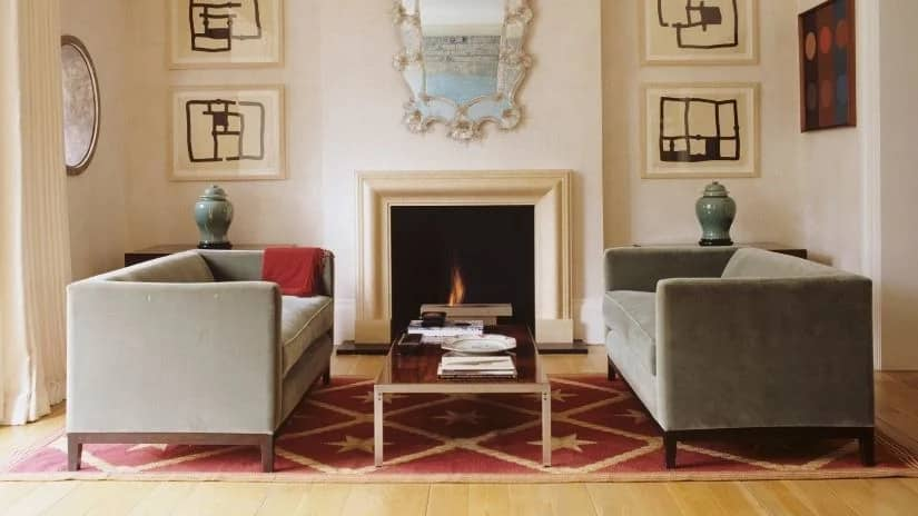 Sala de estar com lareira central de mármore claro e espelho ornamentado acima. Dois sofás baixos em veludo verde-oliva, com linhas retas e bases de madeira escura, estão dispostos simetricamente. No centro, uma mesa de centro baixa em madeira escura com base metálica. A decoração inclui quadros abstratos, vasos de cerâmica verde e um tapete vermelho com padrão estrelado.