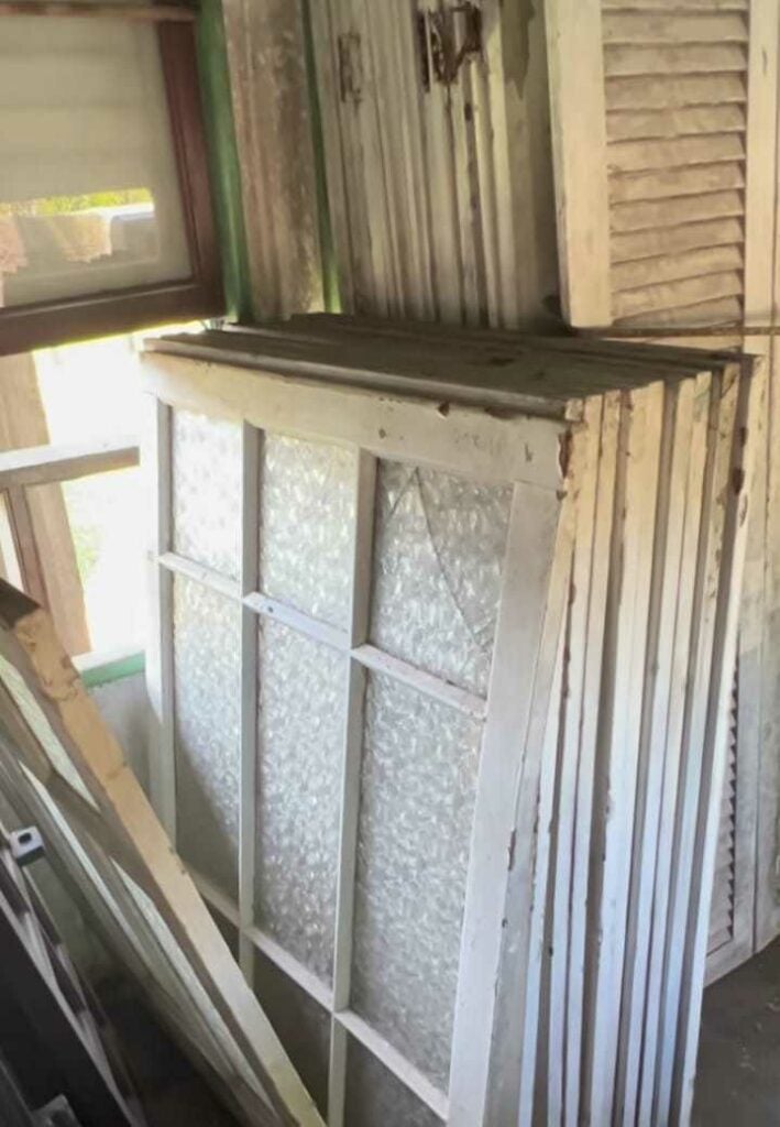Janelas antigas de madeira com vidro texturizado empilhadas em galpão de demolição, prontas para reaproveitamento em projetos criativos.